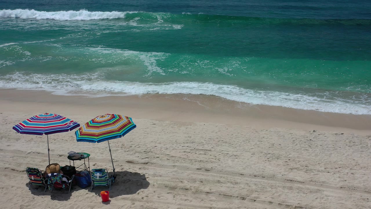 Aerial view in Los Cabos, Mexico with two striped umbrellas and beach gear at the lower left, pale sand across the middle, turquoise surf beyond, and no visible people.
