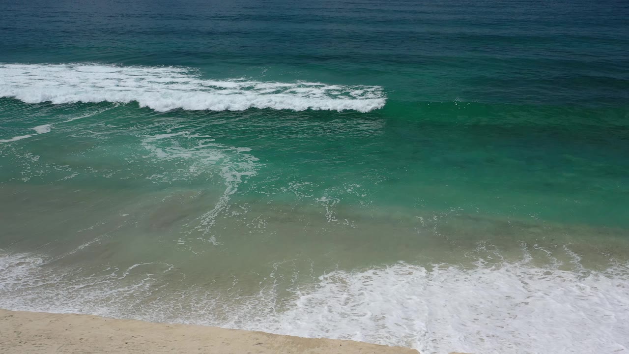 A band of white surf spreads across green shallow water toward pale sand on the Los Cabos shoreline, with no visible people.
