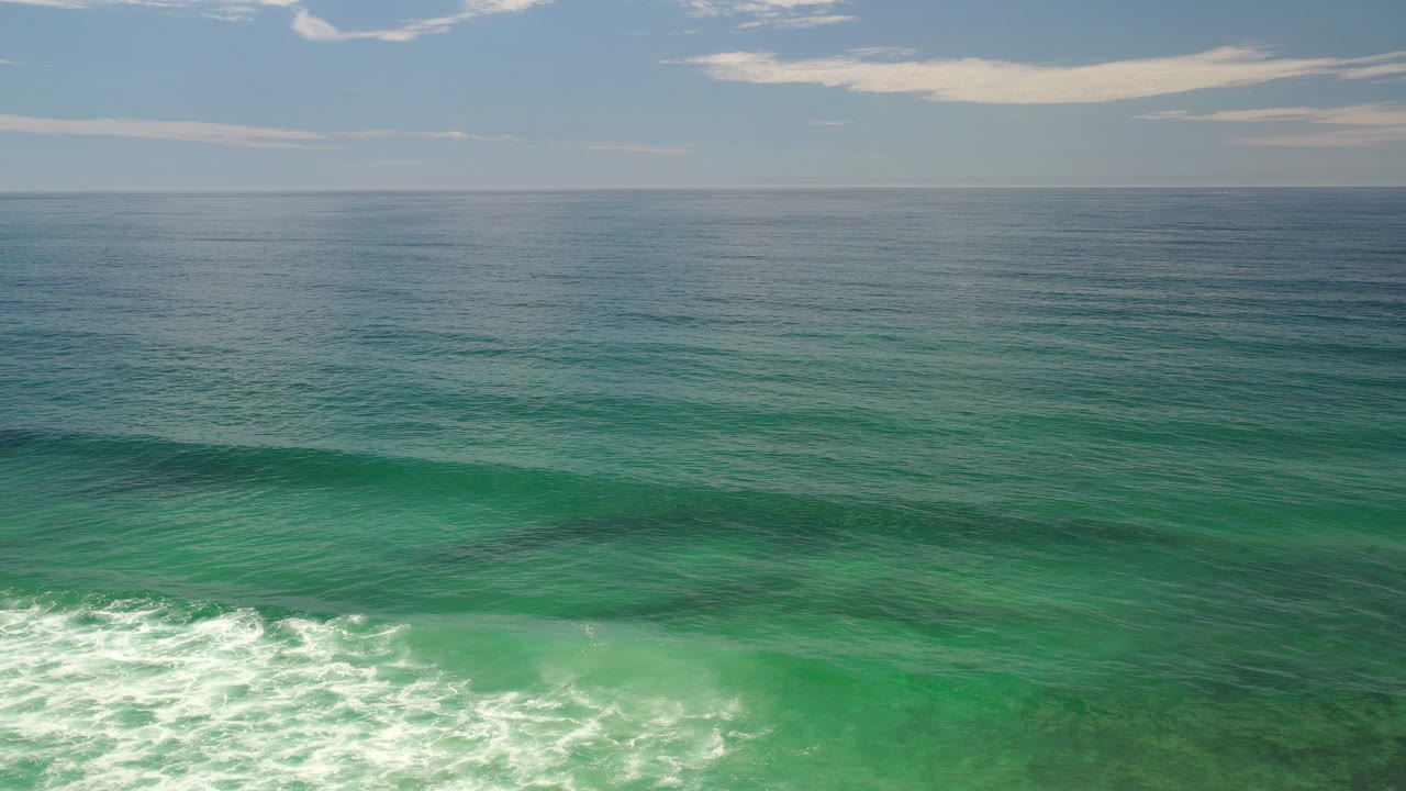 Elevated view of green shallow water in Los Cabos, Mexico, with white surf entering the lower frame, darker seaweed patches below the surface, a distant horizon, scattered clouds, and no visible people.