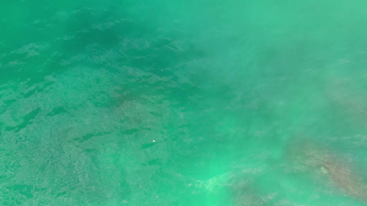 Green-blue Los Cabos water shows darker underwater shapes near the top of frame, tiny sun flecks, and no visible people.