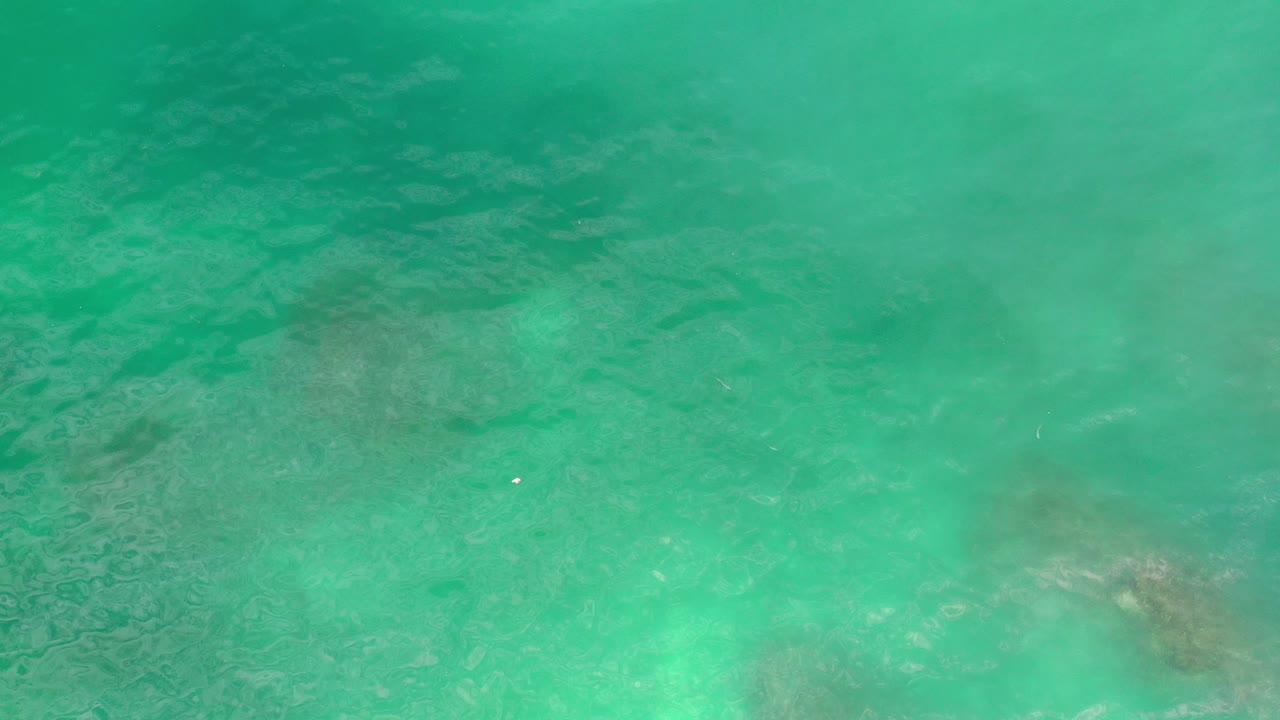 Overhead view of greenish-blue water in Los Cabos, Mexico, with faint ripples, pale cloudy areas below the surface, and no visible people.