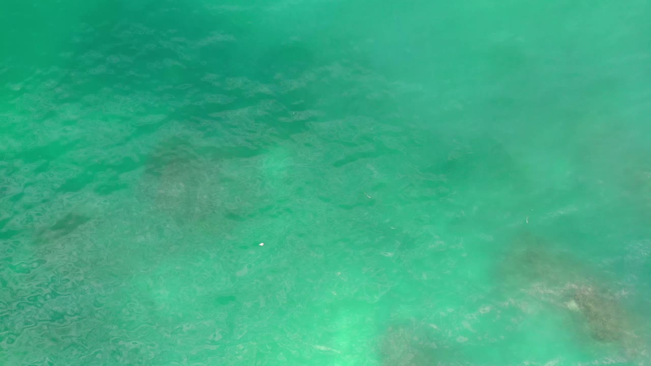 Aerial frame over Los Cabos water showing teal-green surface color, darker submerged patches near the lower half, light ripples, and no visible people.