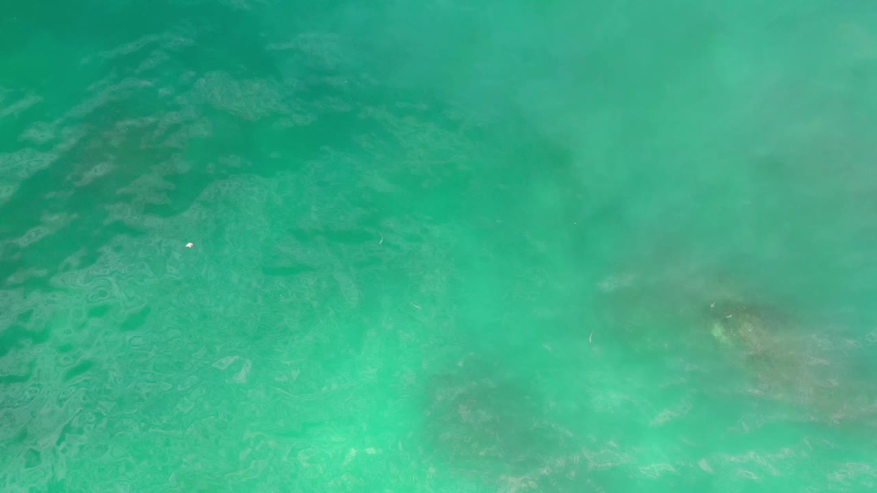 A slightly shifted overhead frame of turquoise Los Cabos water, with soft surface shimmer, muted darker areas, and no visible people.
