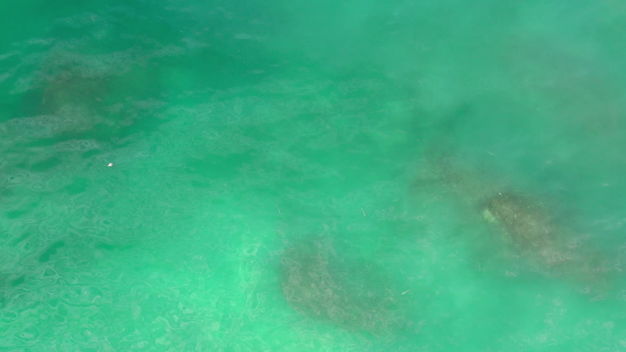 Top-down turquoise seawater with darker submerged rock shapes.