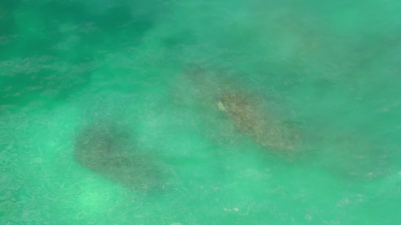Overhead view of green Los Cabos ocean water with darker underwater patches near the upper left and upper right, no visible people.