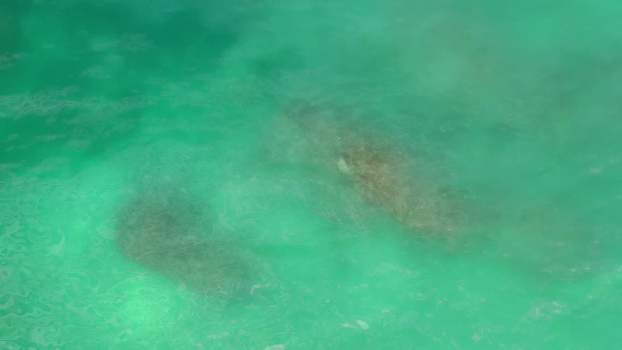 Turquoise Los Cabos water fills the frame as dark submerged rock or reef shapes sit below the rippled surface, no visible people.