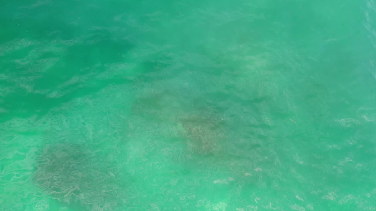 High-angle Los Cabos sea view with turquoise water, soft surface ripples, and muted reef shapes visible beneath, with no visible people.