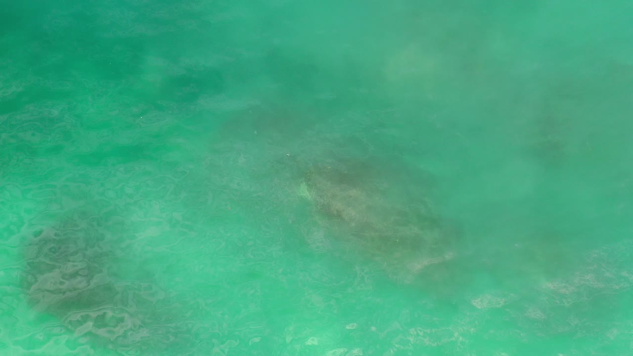 Top-down view of turquoise ocean water in Los Cabos, Mexico, with darker submerged rock patches visible through the surface and no visible people.