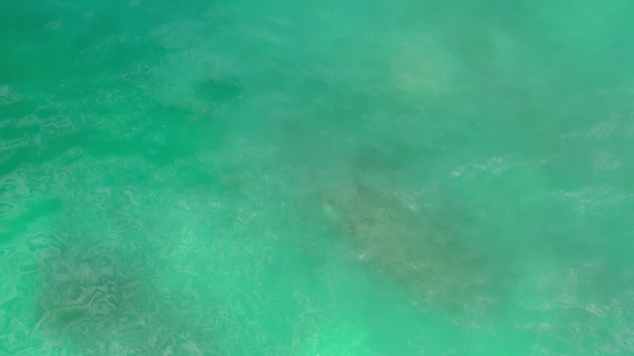 Overhead view of green-blue ocean water in Los Cabos, Mexico, with pale sand and darker underwater patches visible beneath the rippled surface, no visible people.
