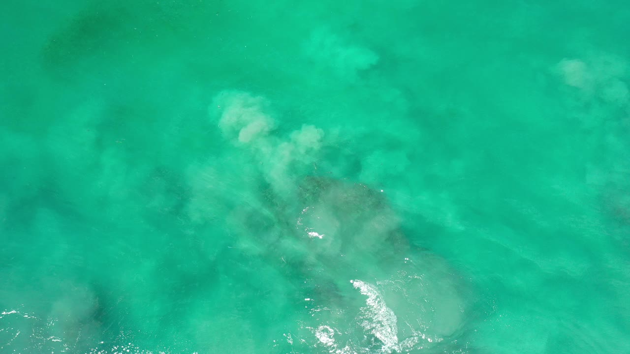 Opening frame of turquoise ocean in Los Cabos, Mexico, with pale underwater clouding, small sun glints, faint dark shapes below, and no visible people.