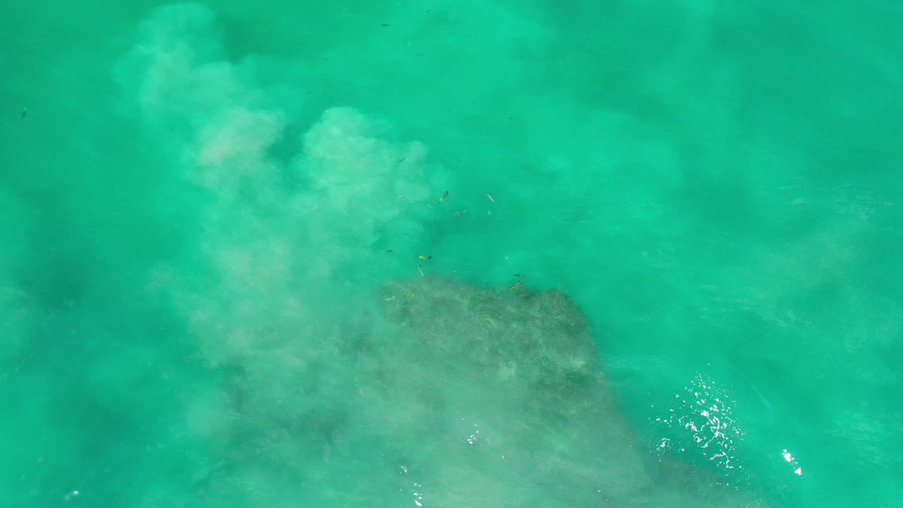 Overhead view of emerald sea water in Los Cabos, Mexico, with pale cloudy wash near the upper left, a darker submerged area below, sun glints on the surface, and no visible people.
