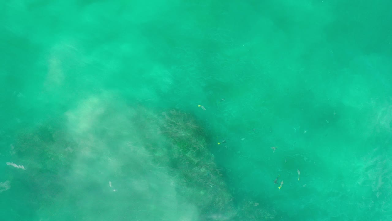 Overhead view in Los Cabos, Mexico of turquoise water with pale clouding over a dark submerged rock patch, with no visible people.
