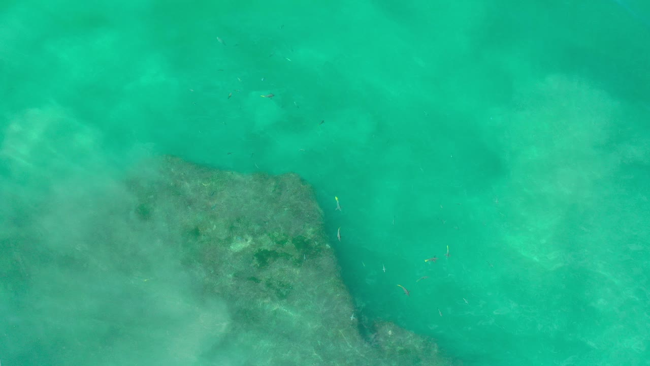 Overhead view of turquoise seawater in Los Cabos, Mexico with a dark submerged rock shelf entering from the lower left and no visible people.