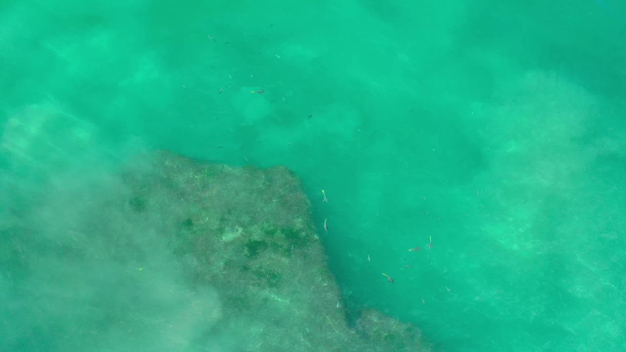 Overhead Los Cabos water frame with green-blue sea, a darker underwater rock patch along the lower edge, faint surface haze, and no visible people.