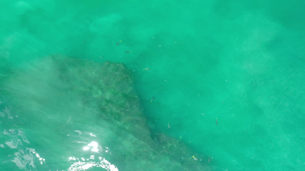 Green ocean water in Los Cabos covers a dark submerged rock near the lower left, with pale cloudy streaks and no visible people.