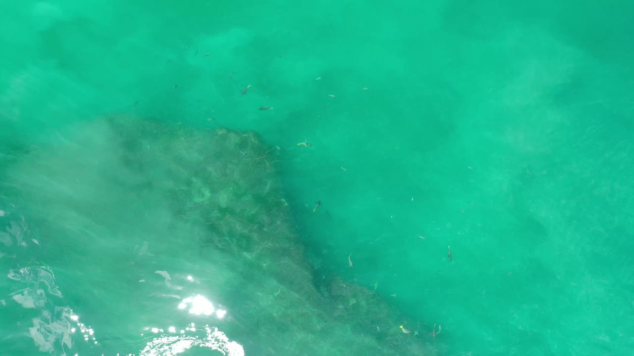 A bright sun glint appears near the lower center as green Los Cabos water passes over the dark rock below, with no visible people.