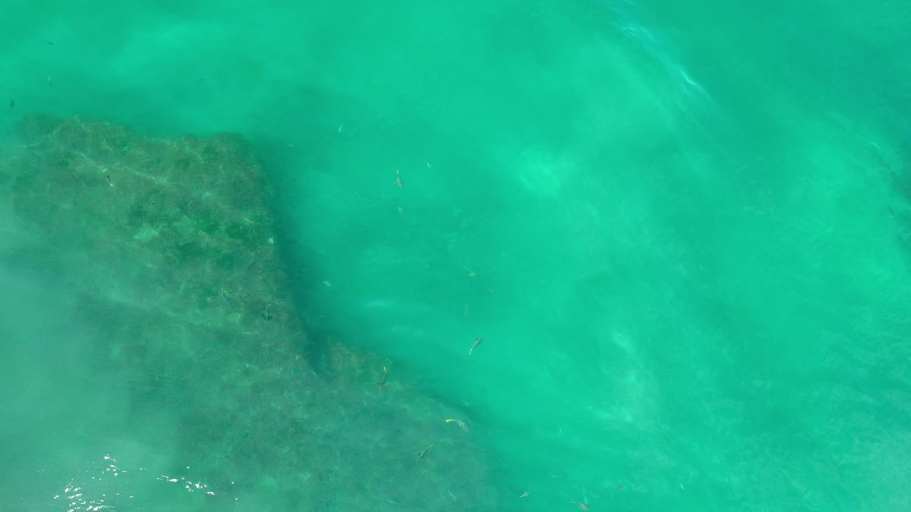 Overhead view in Los Cabos, Mexico of turquoise-green water covering a dark reef patch near the left side, with soft ripples and no visible people.