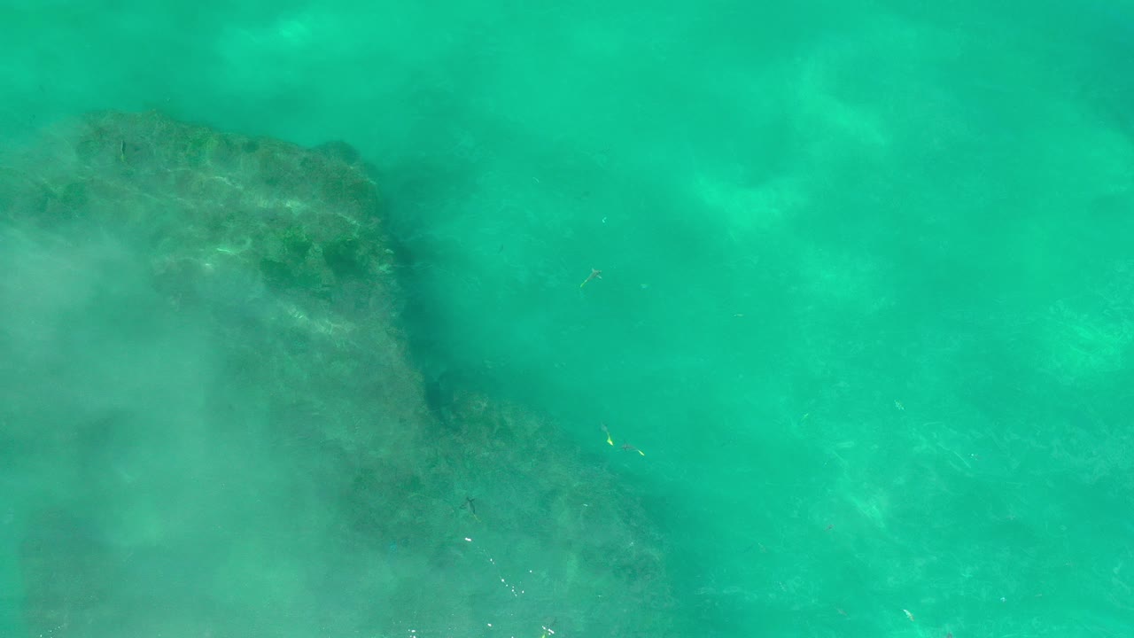 Overhead view of turquoise ocean water in Los Cabos, Mexico, with a dark submerged rock or reef shape entering from the left and no visible people.