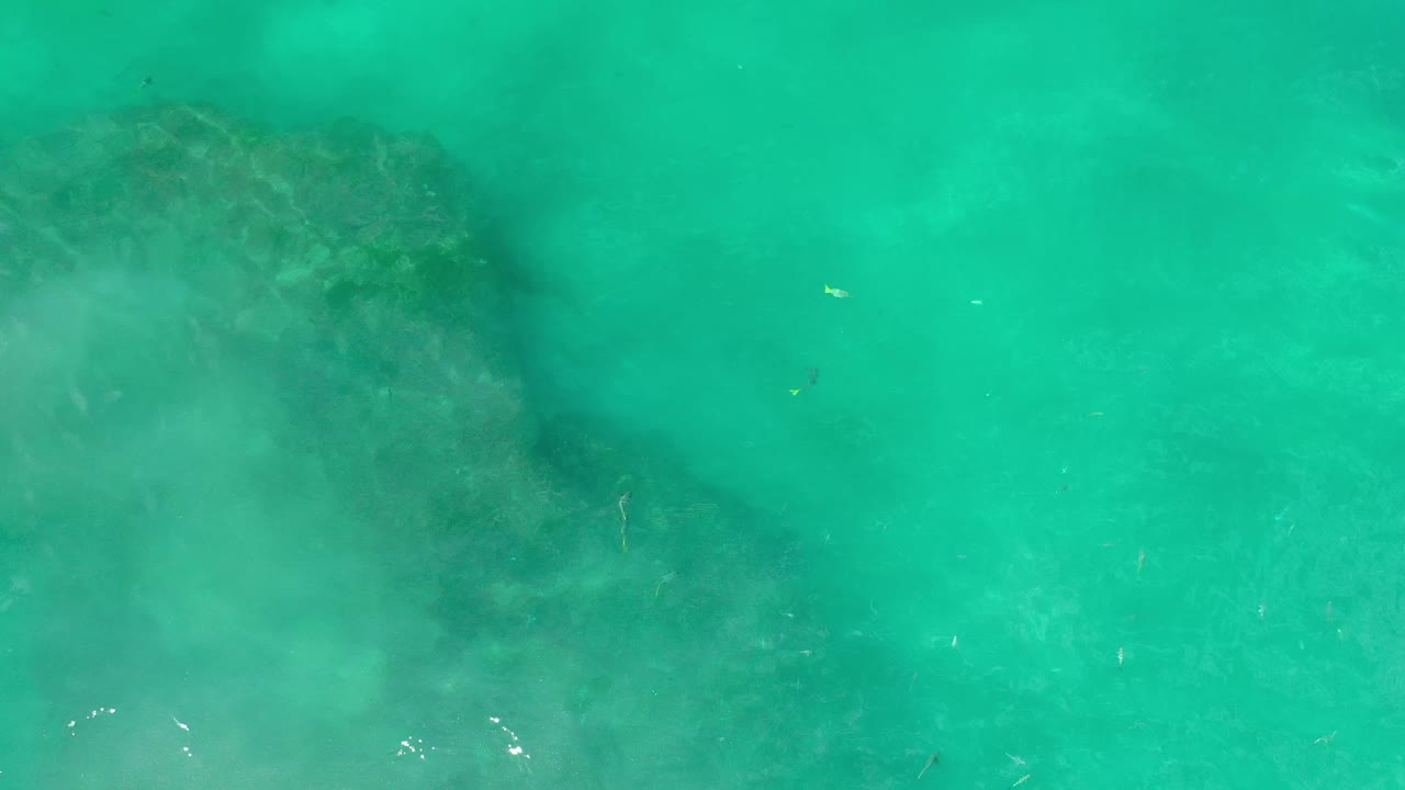 A broad underwater rock shape sits beneath green-blue water in Los Cabos, with soft clouding across the lower frame and no visible people.