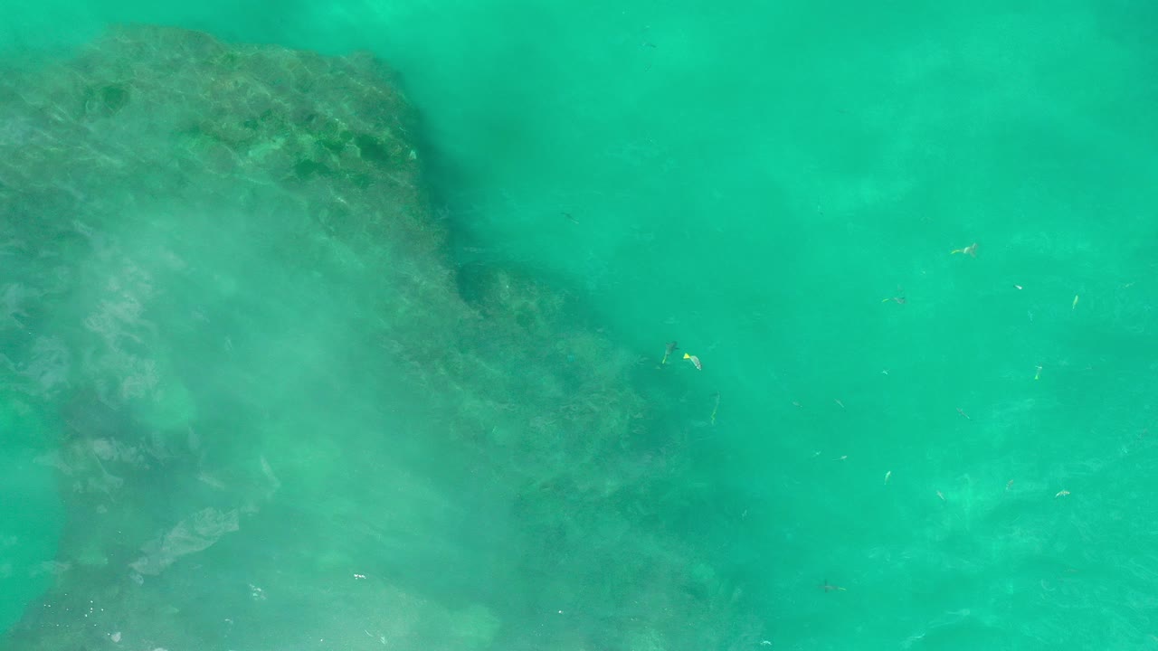 Overhead view of aqua-green ocean water in Los Cabos with a large dark submerged rock shape at left and no visible people.