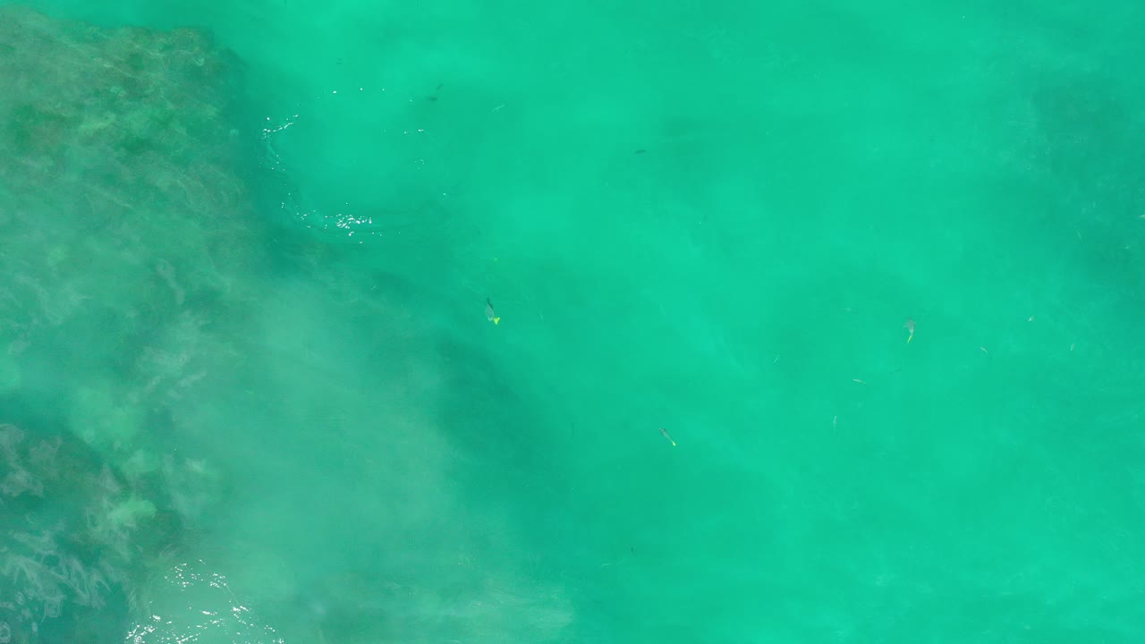 Top-down view of turquoise shallow water in Los Cabos, Mexico, with a dark submerged rock area at upper left and no visible people.