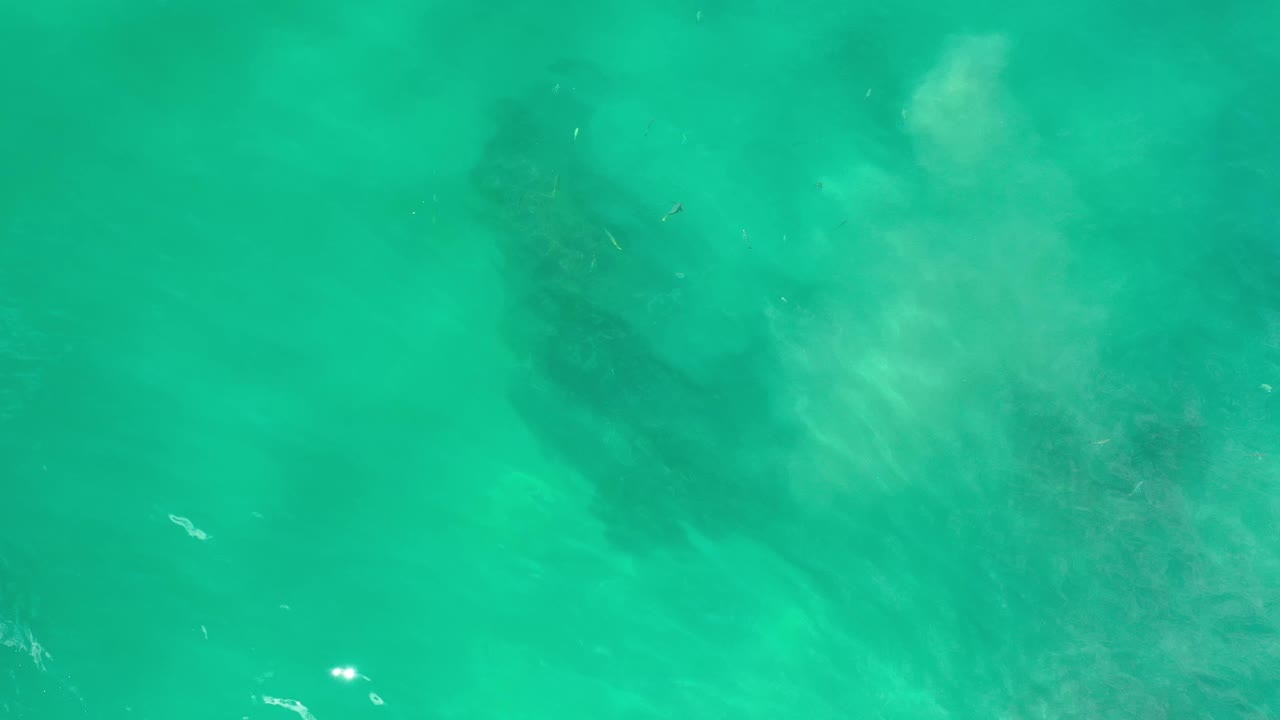 Green Los Cabos seawater viewed from above, with the darker underwater shape centered higher in frame and small white sun glints on the surface, no visible people.