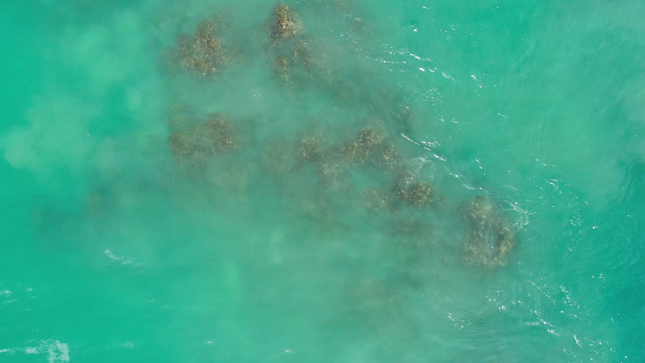 Top-down aerial view in Los Cabos, Mexico, showing turquoise water with several dark submerged rocks near the upper half and no visible people.