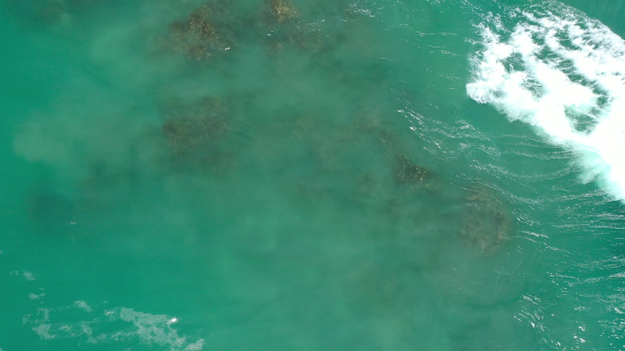 Overhead view in Los Cabos, Mexico, showing turquoise sea over scattered submerged rocks, with a white surf patch entering at the upper right and no visible people.