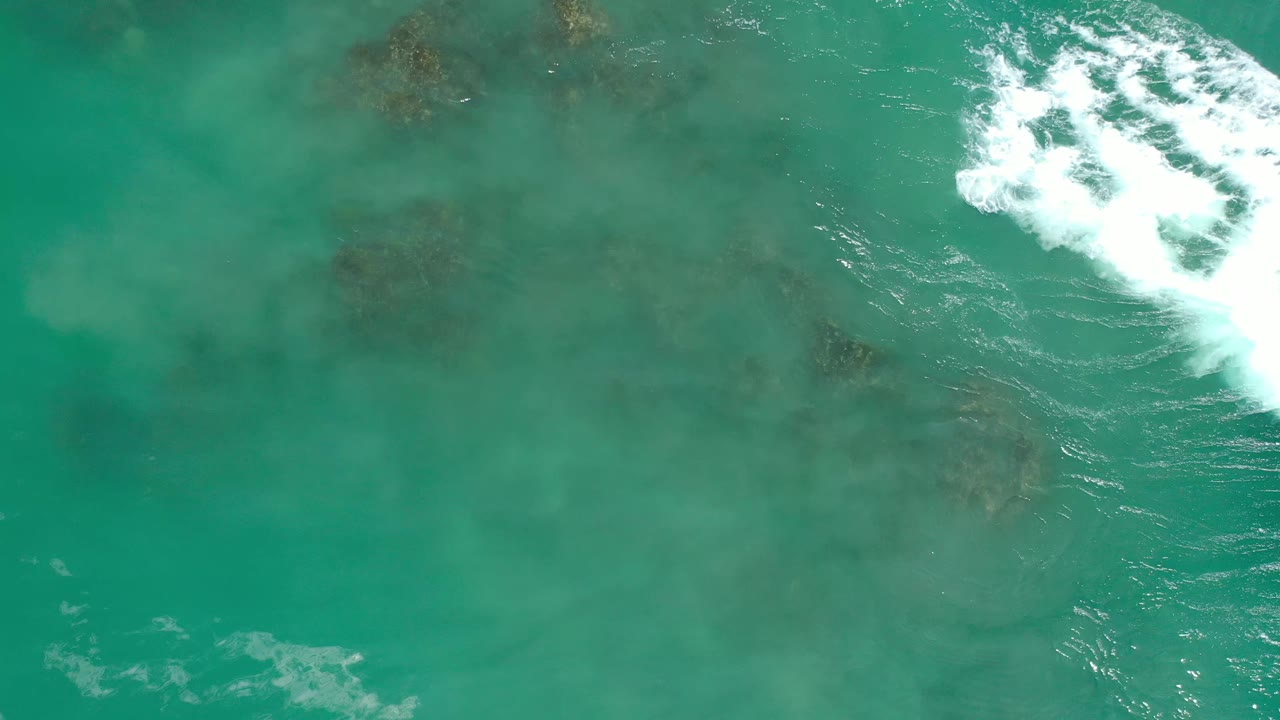 Top-down Los Cabos sea view with faint underwater rock shapes across blue-green water and a bright area of breaking foam along the right edge, with no visible people.