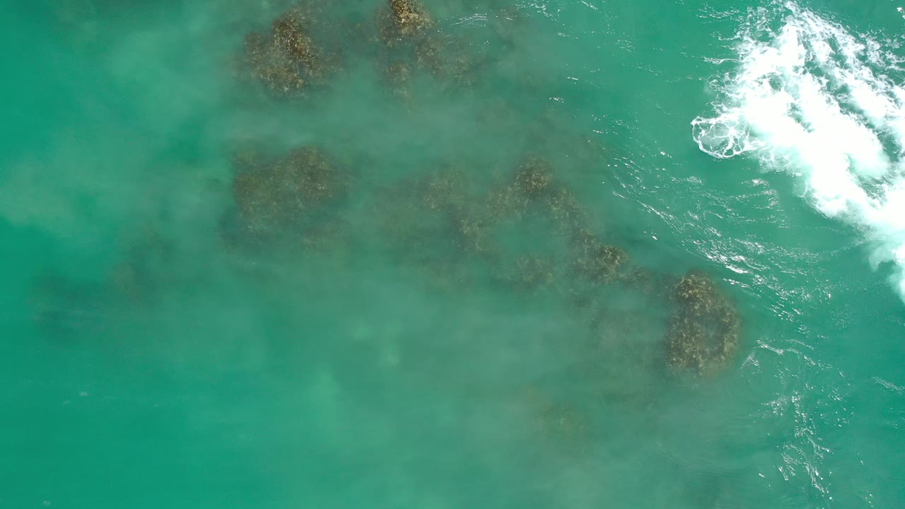 Aerial sea texture with several dark rocks below water and surf at top right