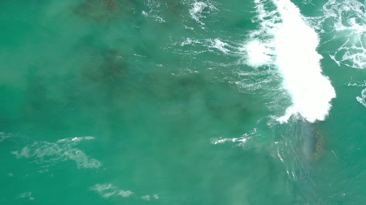 Overhead-looking view of turquoise water in Los Cabos, Mexico, with a bright white wave breaking over a darker submerged reef patch and no visible people.