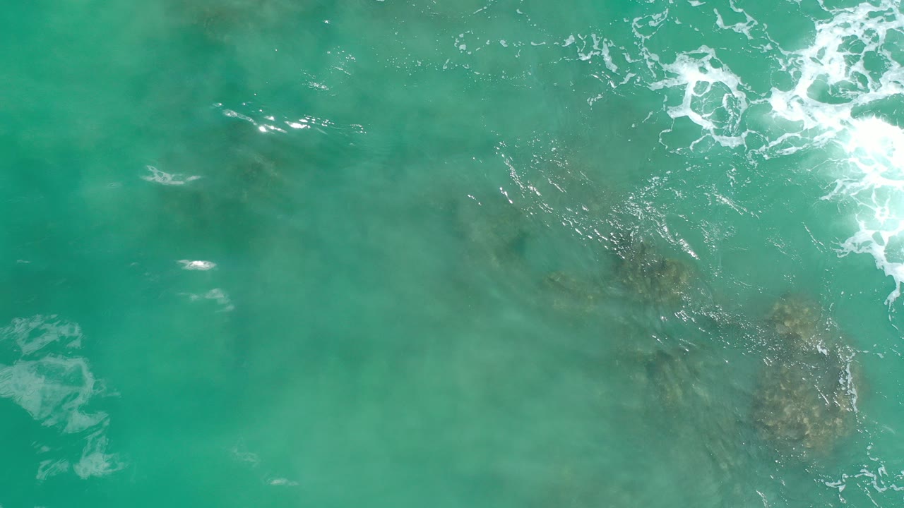 Overhead view of turquoise water in Los Cabos, Mexico, with white foam near the upper right and dark submerged patches below the surface, no visible people.
