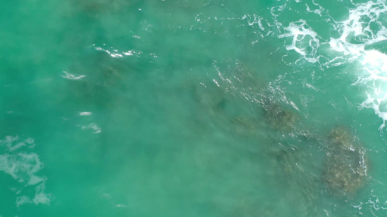 Aerial frame over green-blue Los Cabos water with a darker underwater area near center right and small foam marks across the surface, no visible people.