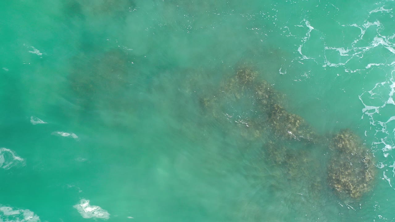 Overhead view of turquoise ocean water in Los Cabos, Mexico, with dark submerged rock or reef patches near the center and no visible people.