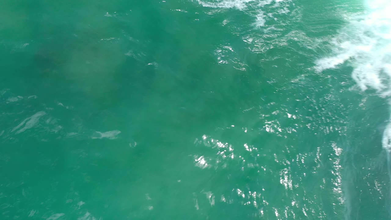 Overhead Los Cabos water frame with bright sun reflections, soft foam at the top right, and a pale submerged streak in the center, no visible people.