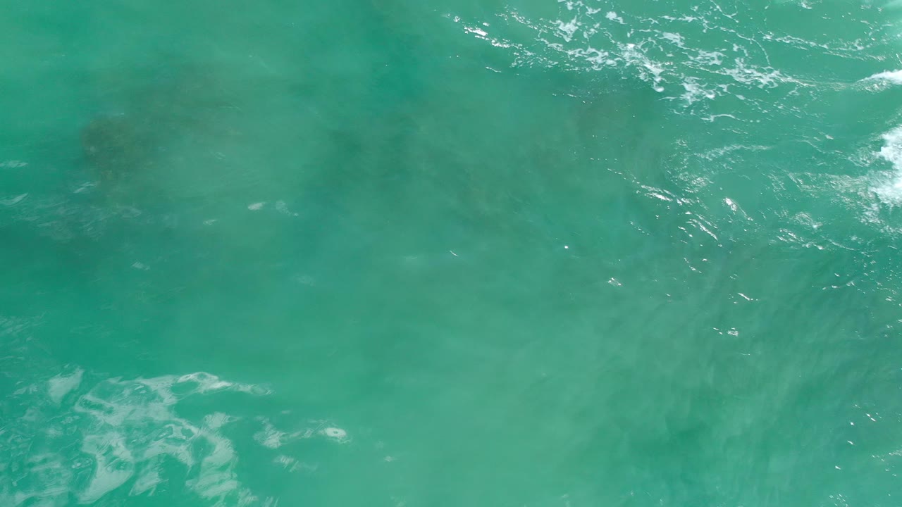 Overhead view in Los Cabos showing smoother aquamarine water, faint cloudlike underwater color changes, and thin white foam along the upper edge, no visible people.