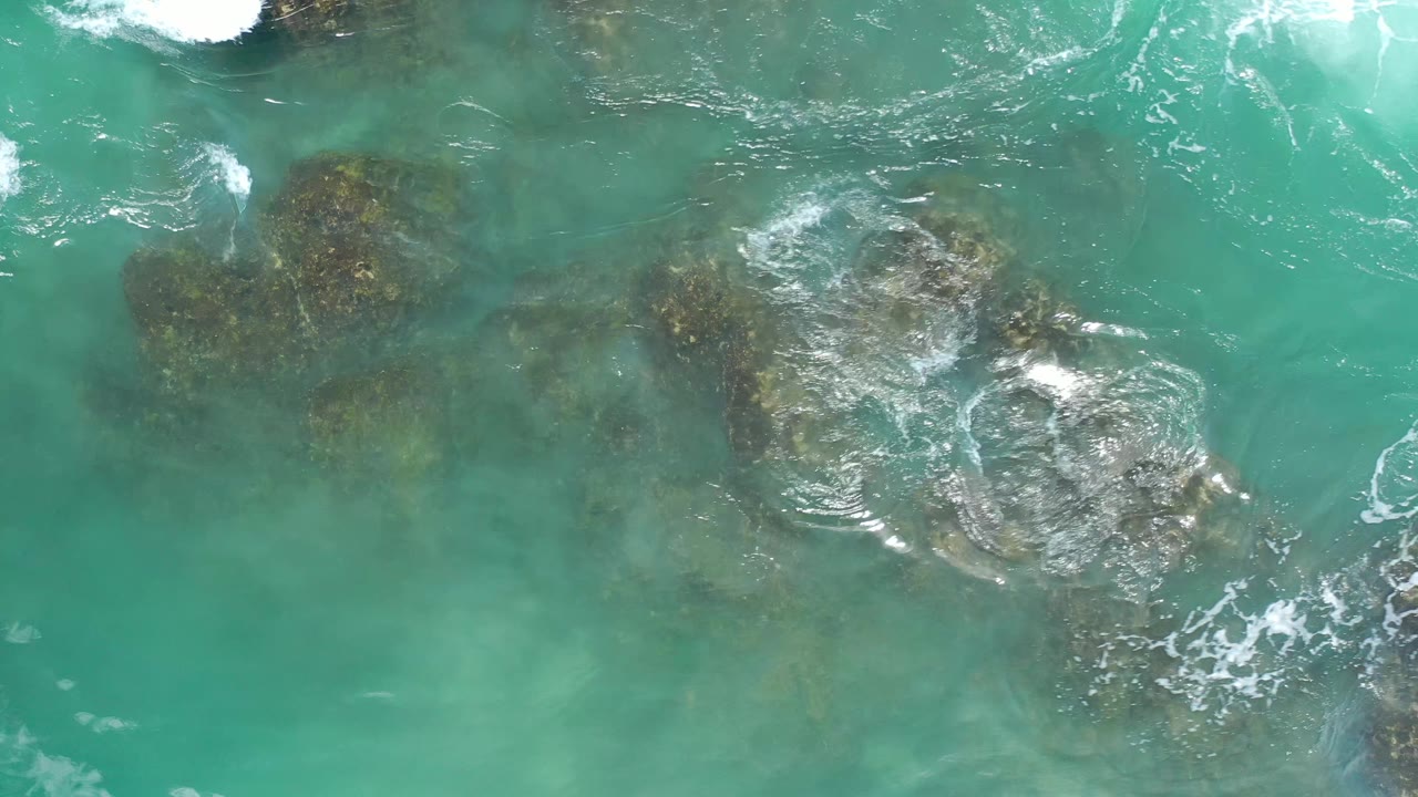 Overhead frame of blue-green ocean water in Los Cabos, Mexico, with foam curling around a central rock cluster and no visible people.
