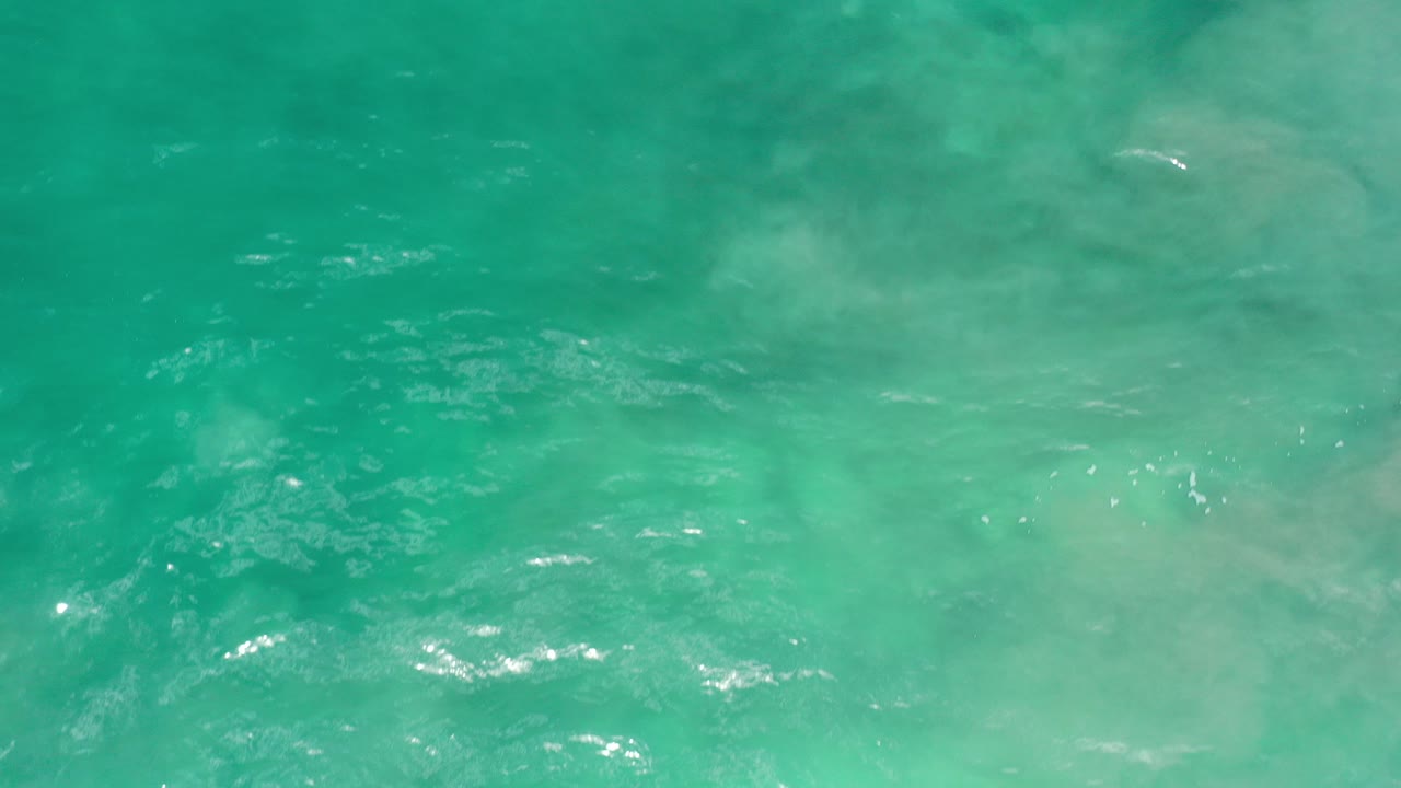 Overhead-looking view of green-turquoise water in Los Cabos, Mexico, with pale underwater clouding, soft ripples, scattered sun glints, and no visible people.
