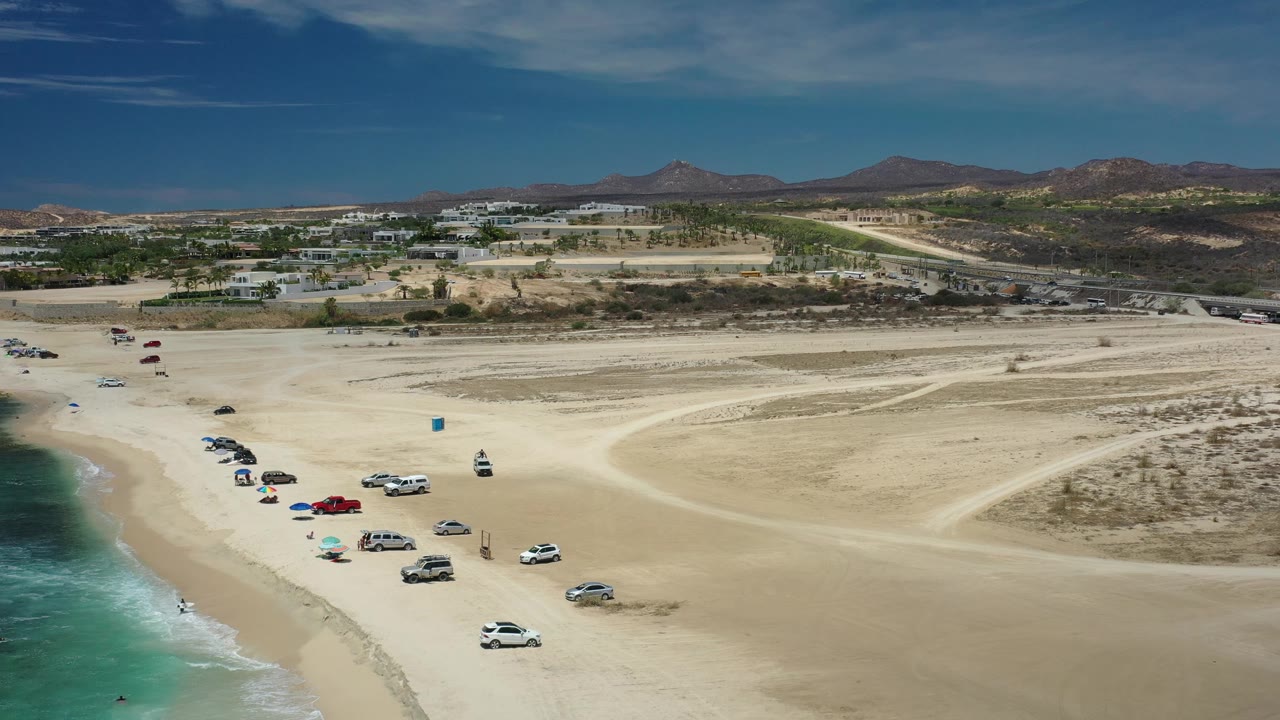 Los Cabos beach aerial with the surf strip at left, vehicles parked near umbrellas, open sandy flats to the right, and low development backed by hills.