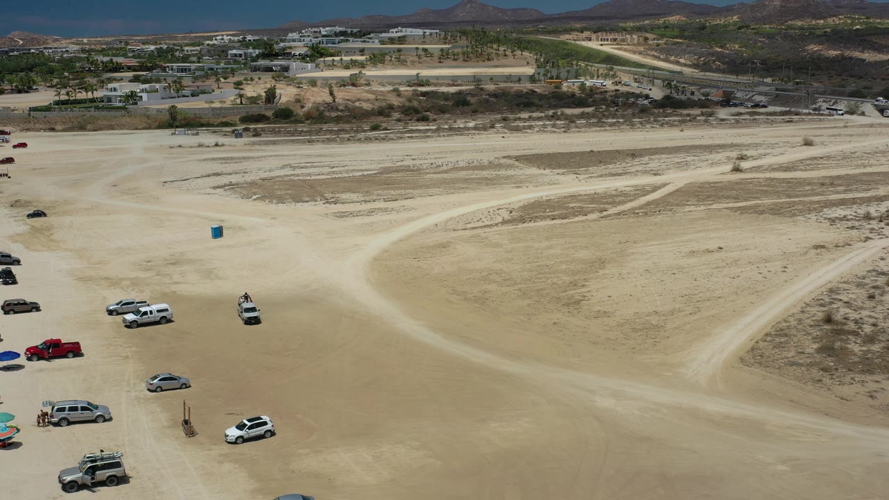 Aerial Los Cabos frame with a red pickup, white SUVs, and other cars parked on sand, plus a small blue unit and pale vehicle tracks crossing the open ground.