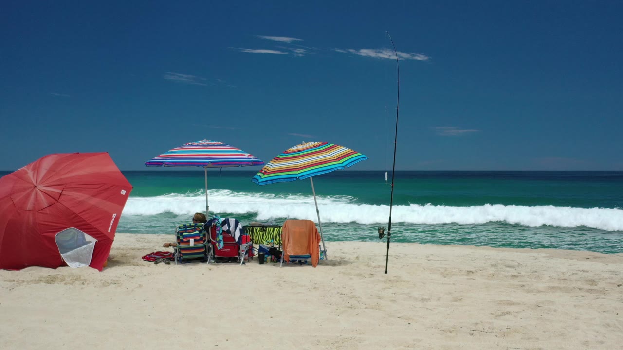 A Los Cabos shoreline view shows beach chairs clustered beneath striped umbrellas, people partly visible in the shade, and a white wave line rising behind them.