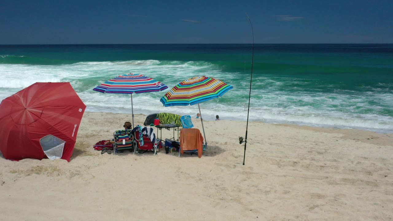 Los Cabos shoreline from above with a red umbrella, two colorful striped umbrellas, seated beachgoers, packed gear, and teal water rolling toward the sand.