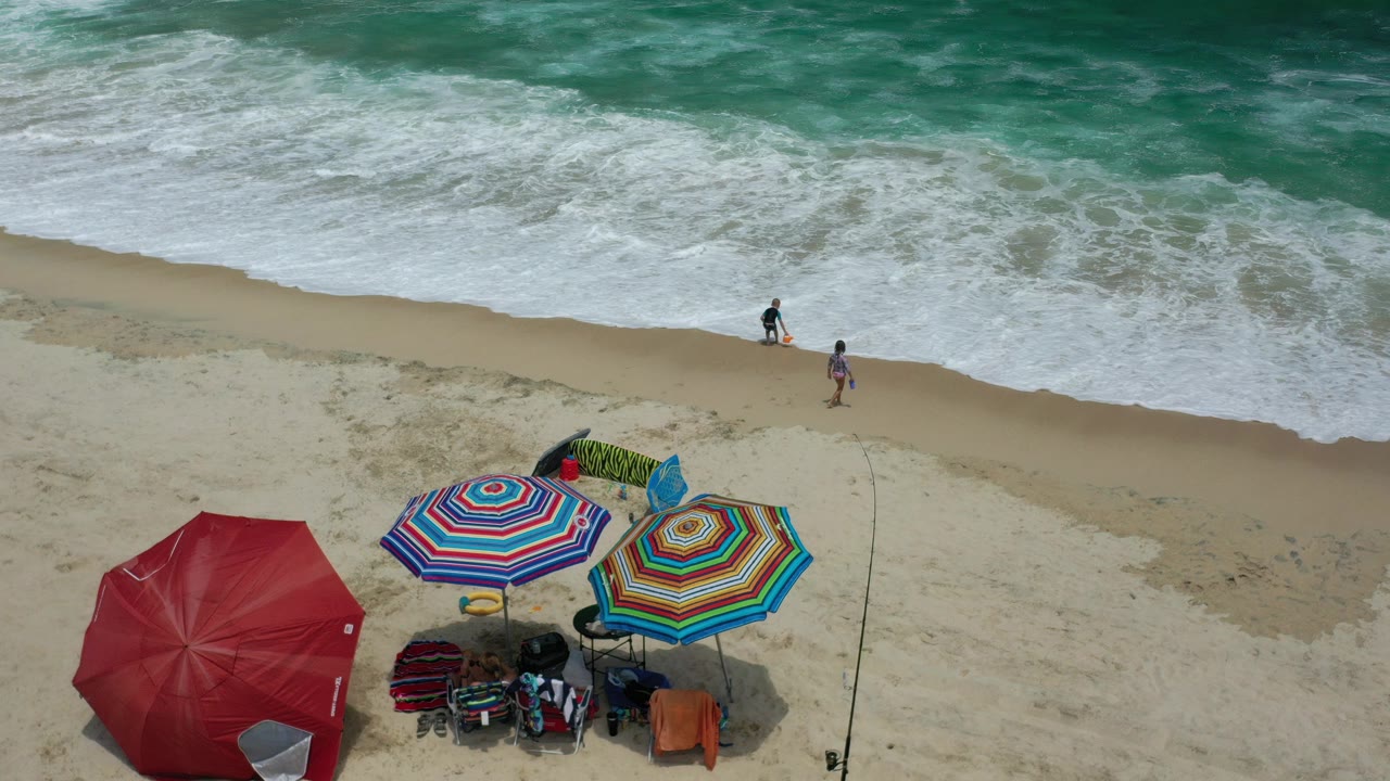 Overhead beach view with umbrellas, surf, and two people near the waterline.