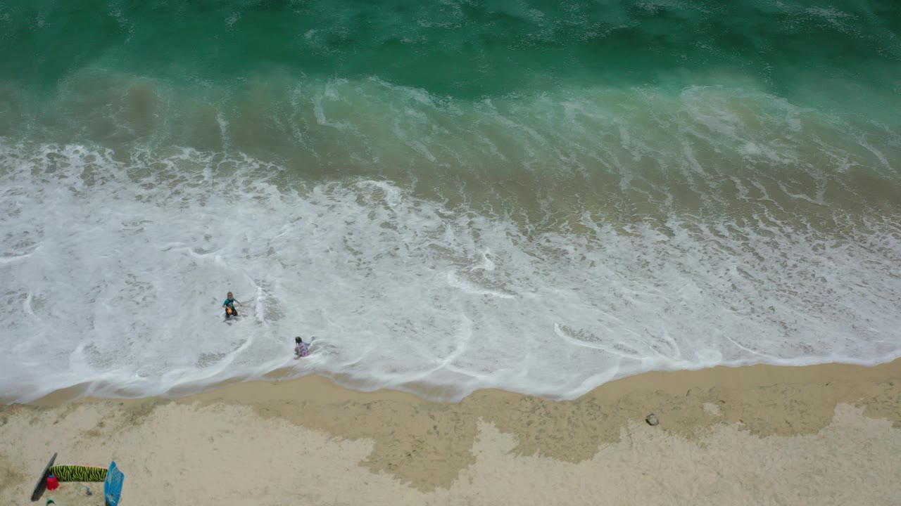 Overhead view in Los Cabos, Mexico, showing white surf spreading across tan sand with two small people near the edge of the waves and beach gear partly visible below.