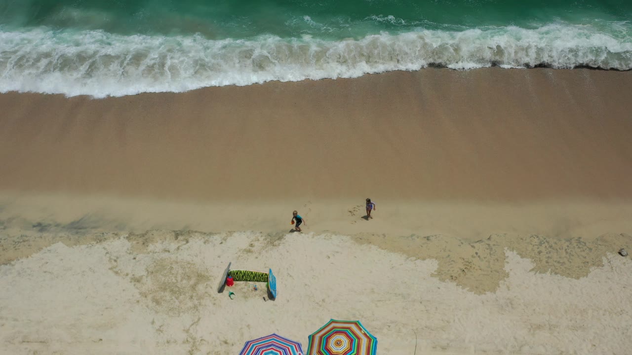 Top-down view of a Los Cabos beach with green-blue surf at the top, pale sand below, small people on the beach, and striped umbrellas partly visible at the bottom.