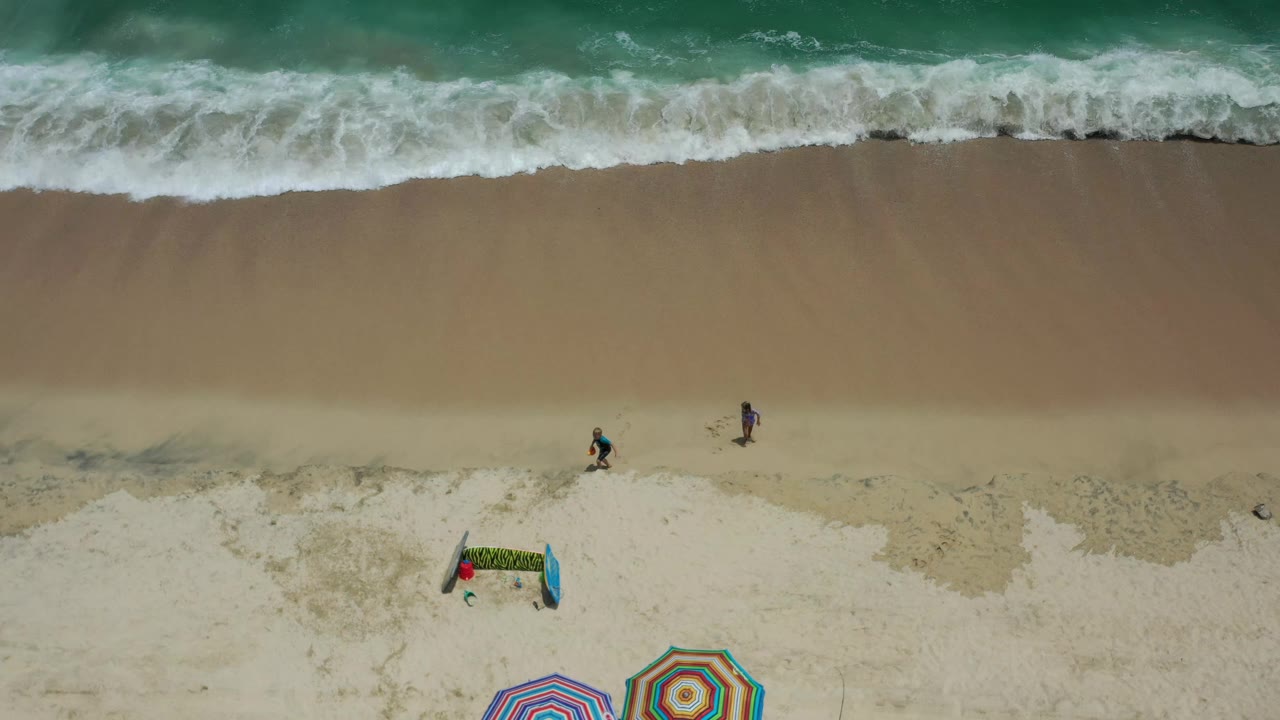 Overhead Los Cabos beach frame with the breaking wave line across the top, two small figures on the sand, and colorful striped umbrellas entering from the bottom edge.