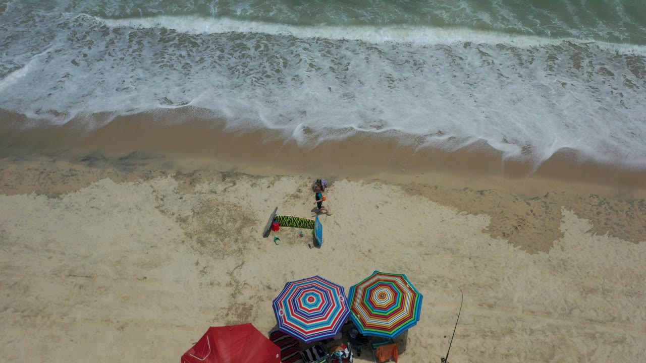 Overhead beach view with surf, umbrellas, fishing rod, and children near sand toys.