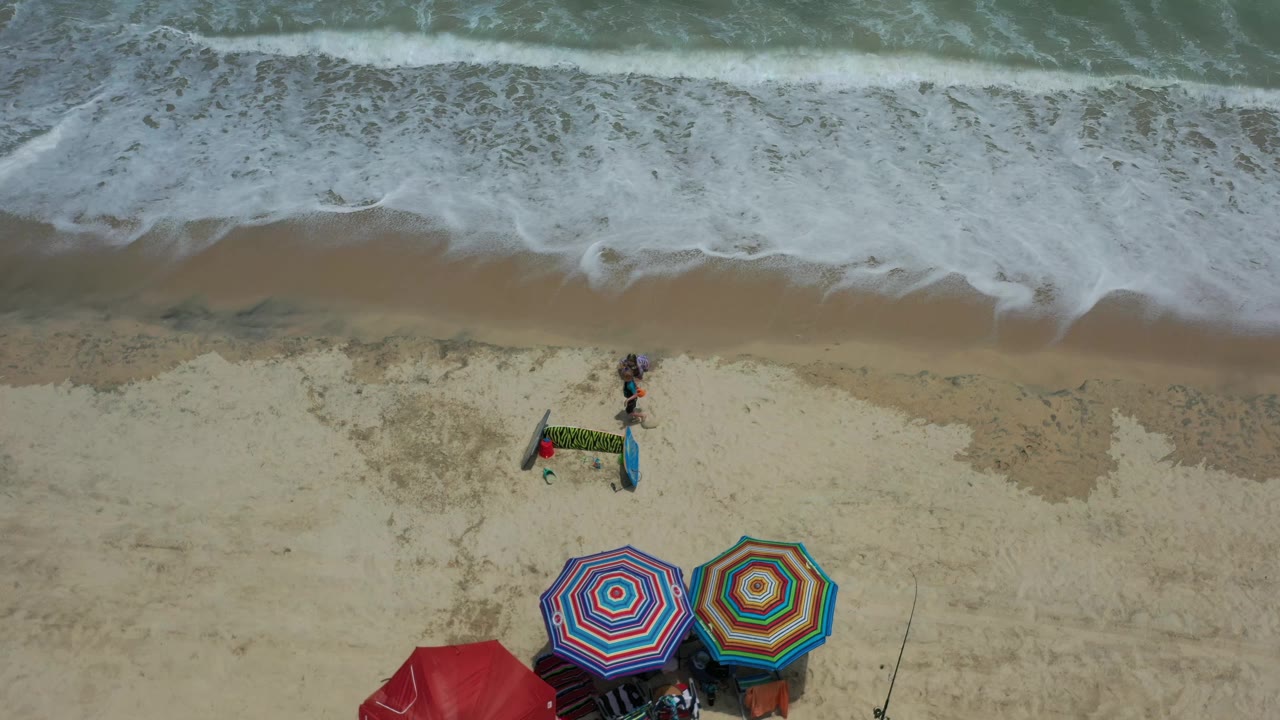 Overhead Los Cabos beach frame with the surf line high in the image, two striped umbrellas low in frame, and children visible beside a small play area on the sand.