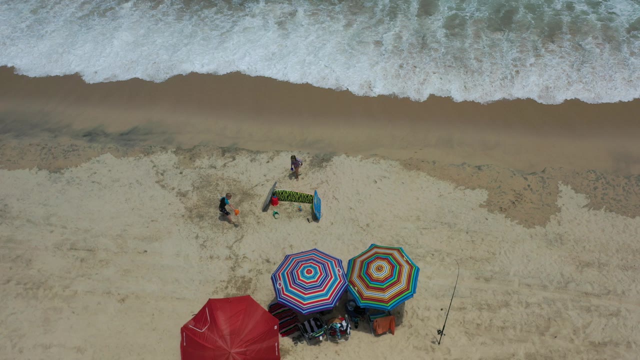 Overhead beach view with surf, umbrellas, and children near sand toys.