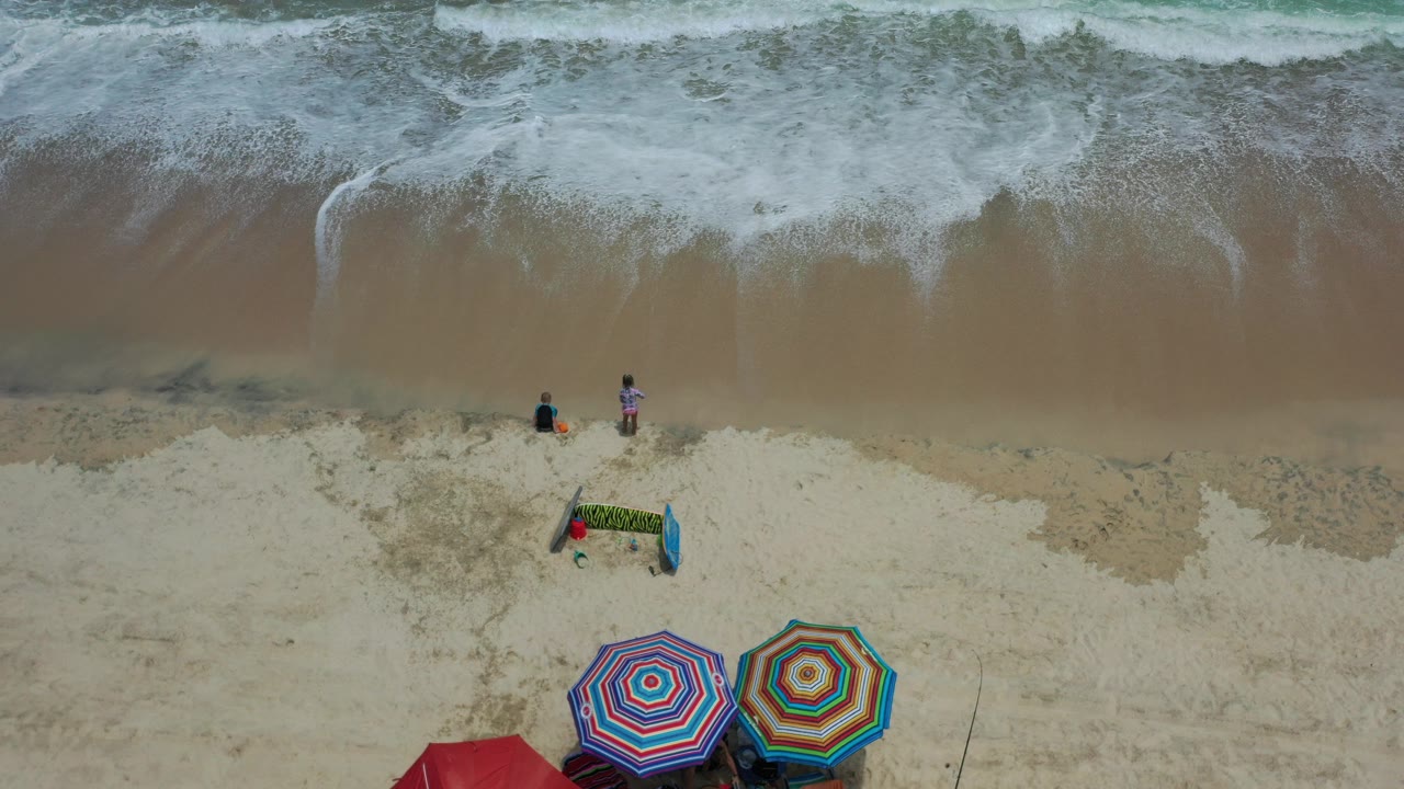 Overhead frame in Los Cabos, Mexico showing two children near the waterline, foamy surf at the top, pale sand, and colorful umbrellas along the lower edge.